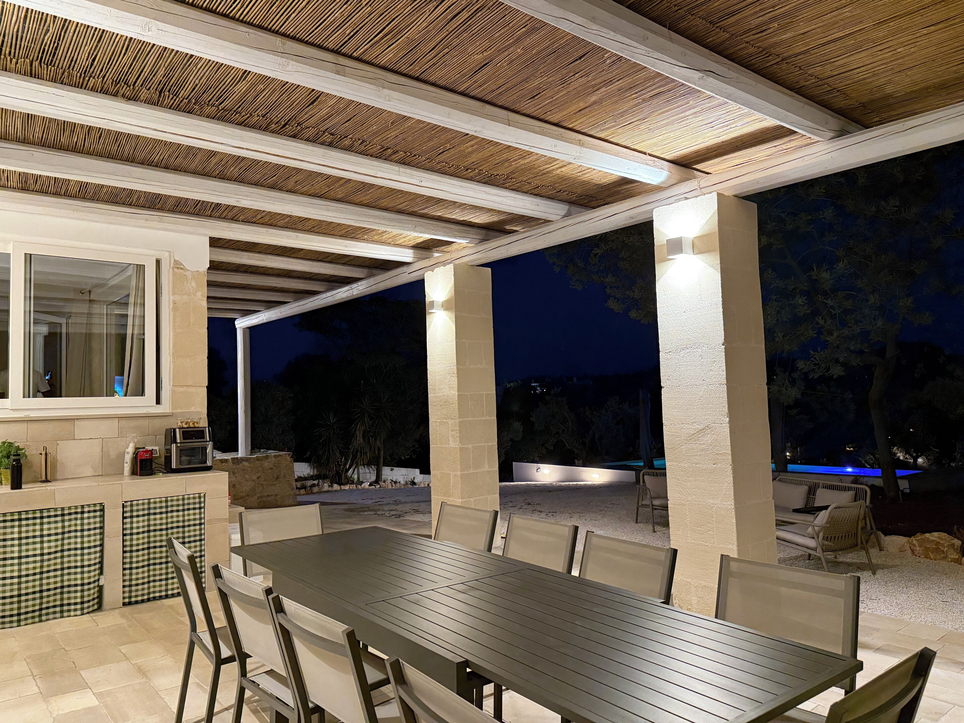 Un patio exterior con mesa y sillas, una parrilla incorporada y una piscina al fondo.