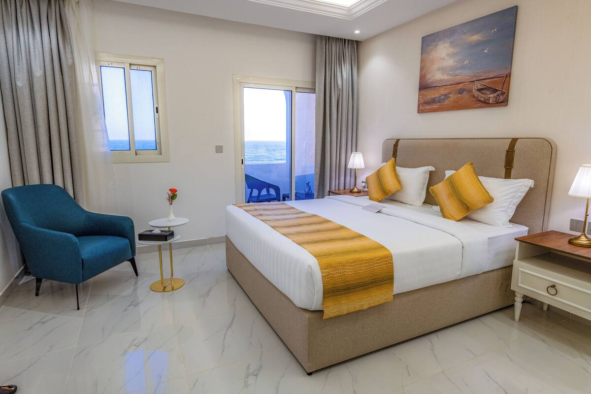 Habitación de hotel con una cama grande, un sillón azul, una mesita con un florero y un cuadro en la pared.