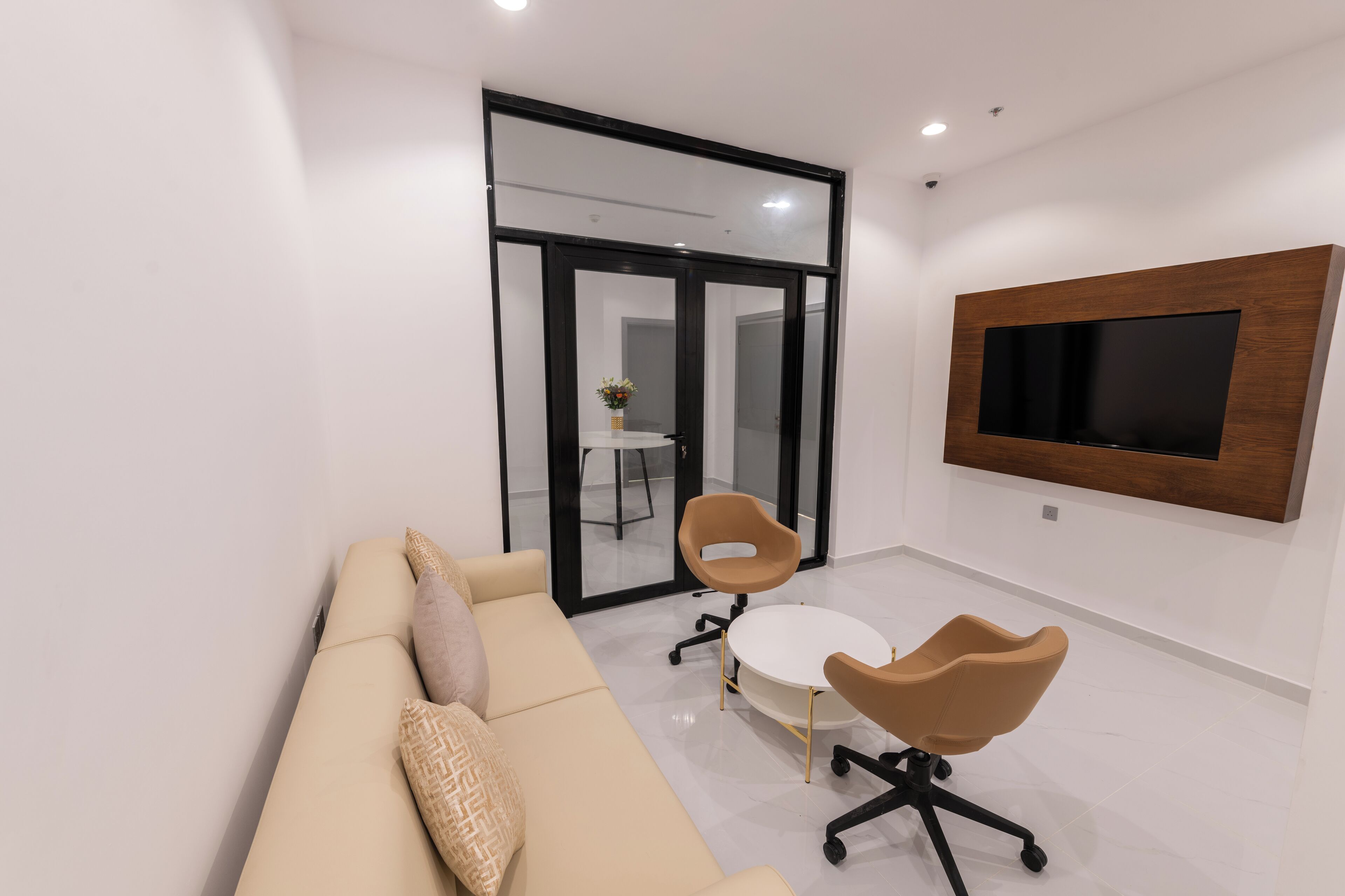 A modern office space with a beige sofa, two brown chairs, a white table, and a mounted TV.