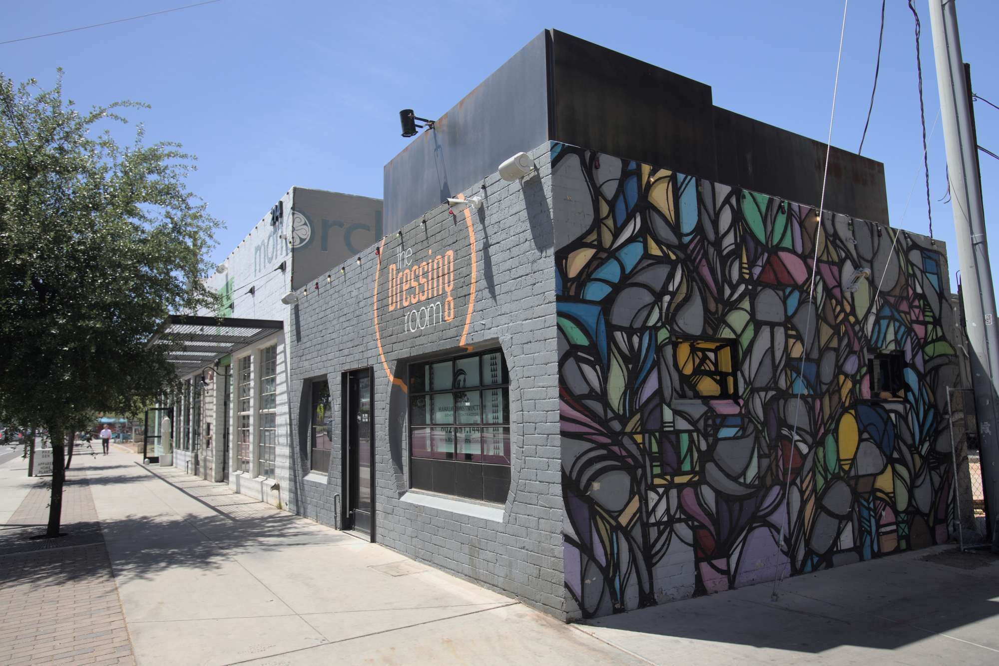 A building with a colorful mural on the side, a storefront with a sign reading "The Dressing Room," and a clear sky above.
