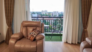 Una sala de estar con un sofá de cuero, un cojín decorativo estampado, una mesa de comedor con sillas y vistas a un paisaje urbano a través de amplios ventanales.