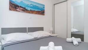 Een hotelkamer met een bed, witte kussens, een grijze plaid en een schilderij van een berglandschap aan de muur.