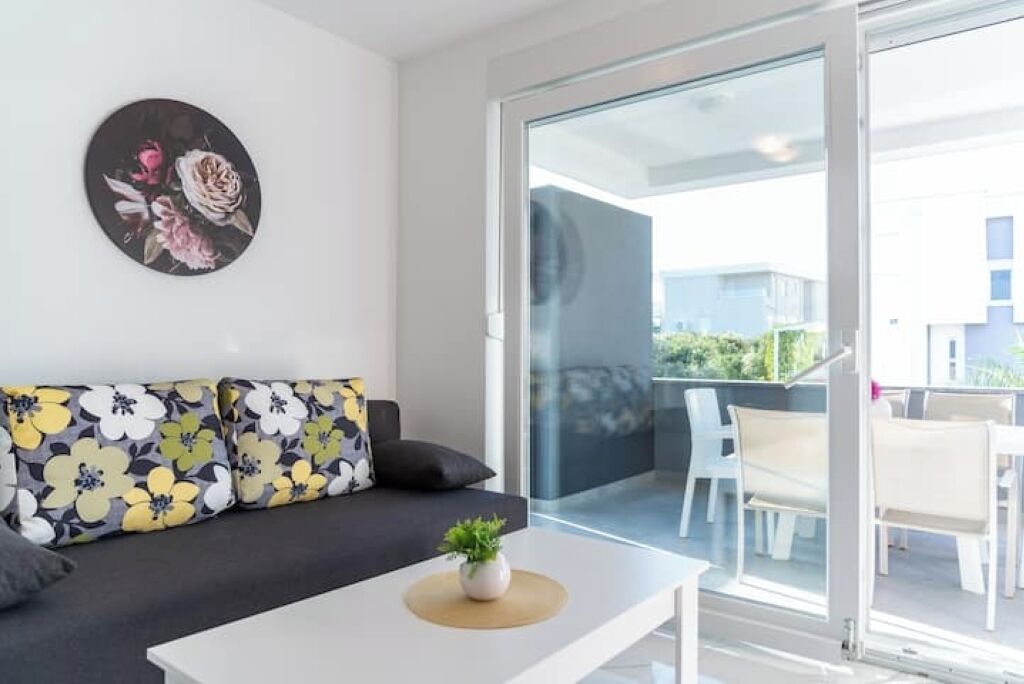 Ein modernes Wohnzimmer mit einer schwarzen Couch, einem weißen Couchtisch und einer Glastür, die auf einen Balkon mit Outdoor-Möbeln führt.