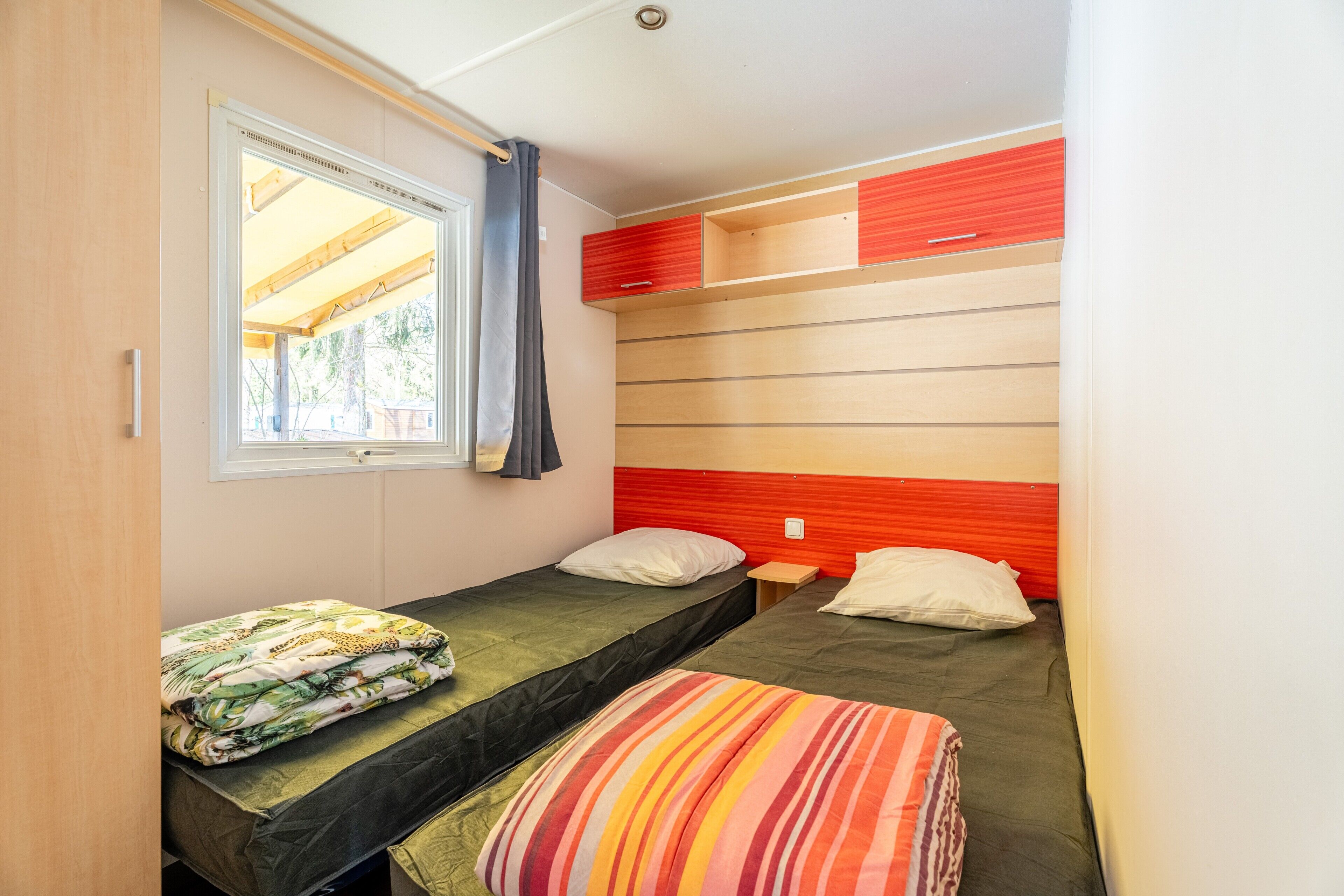 A compact room with two single beds, a small table, a window with curtains, and a red and beige headboard.