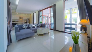 A modern living room with a grey sofa, a coffee table, and a view of the kitchen and outdoor area through large glass doors. - Beautiful 4-Bedroom Pool Villa BL9 (Hua Hin)