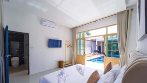 A hotel room with a bed, a TV, an air conditioning unit, a bathroom with a shower, and a view of an outdoor swimming pool. - Beautiful 4-Bedroom Pool Villa BL9 (Hua Hin)