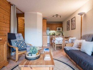 A cozy living room with a wooden chair, a sofa, a dining table, and a kitchen in the background. - Close to Ski Piste Prarion - Swimming pool & Gym (Les Houches)