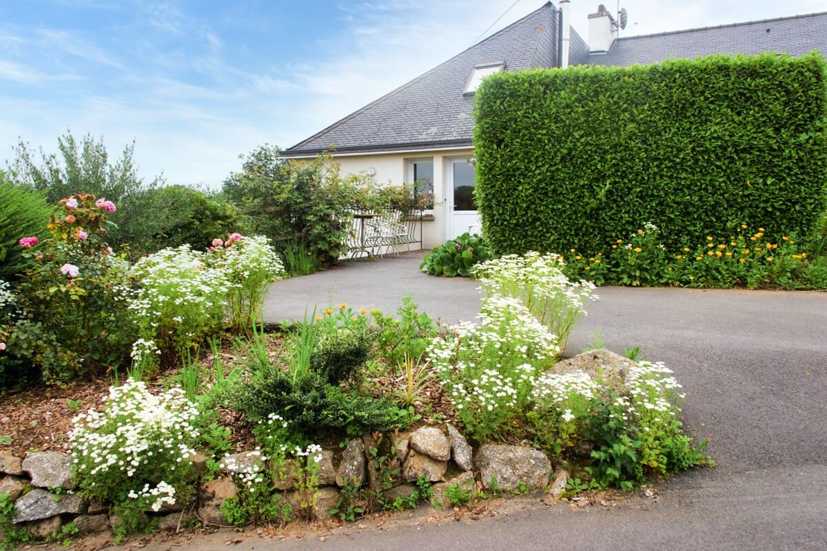 Ein Wohnhaus mit befestigter Auffahrt, einer Steinmauer und verschiedenen blühenden Pflanzen.