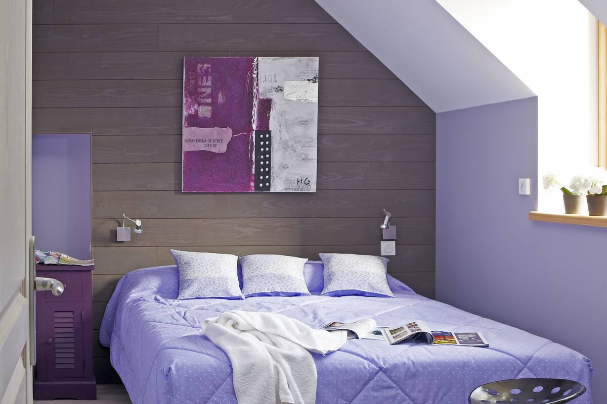 Ein Schlafzimmer mit einem großen Bett, einem hölzernen Kopfteil, einer violetten Wand und einem kleinen Fenster mit Ablage.