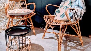 Un rincón acogedor con sillas de mimbre y una cesta tejida, un taburete tapizado de color negro, y vistas a una zona de bar a través de un separador de vidrio.
