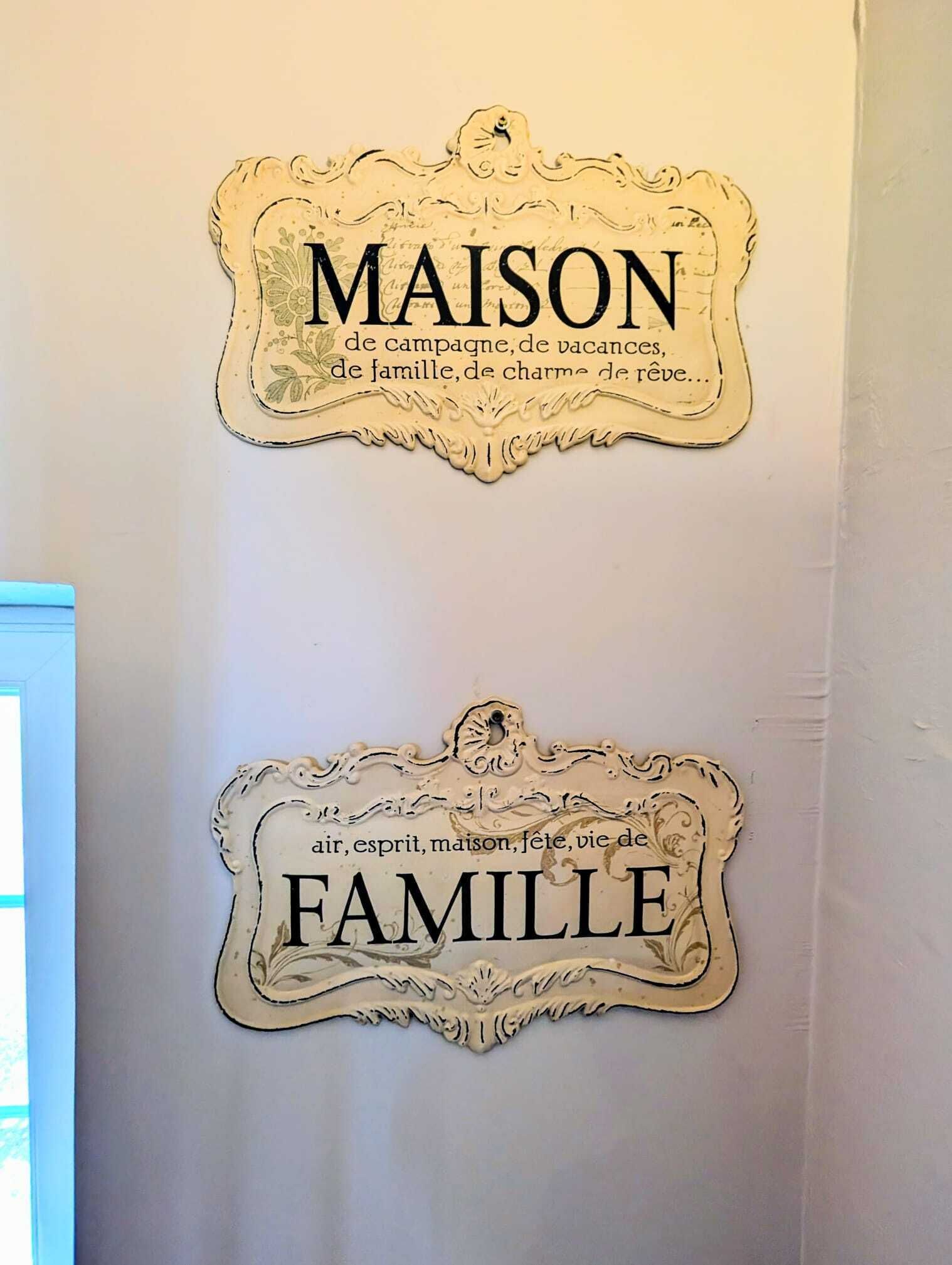 Two decorative signs on a wall, one labeled 'MAISON' and the other 'FAMILLE', with ornate designs and text indicating themes of home and family.