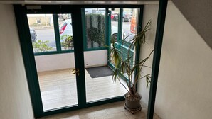 A view from a room with a sliding glass door leading to a balcony. The balcony has a wooden floor and is bordered by a green railing. There is a potted plant on the balcony floor.