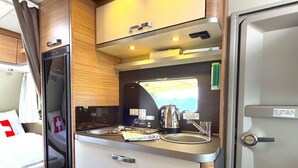 A compact kitchenette with a stove, sink, and cabinets, a bed with a window, and a door with a handle.