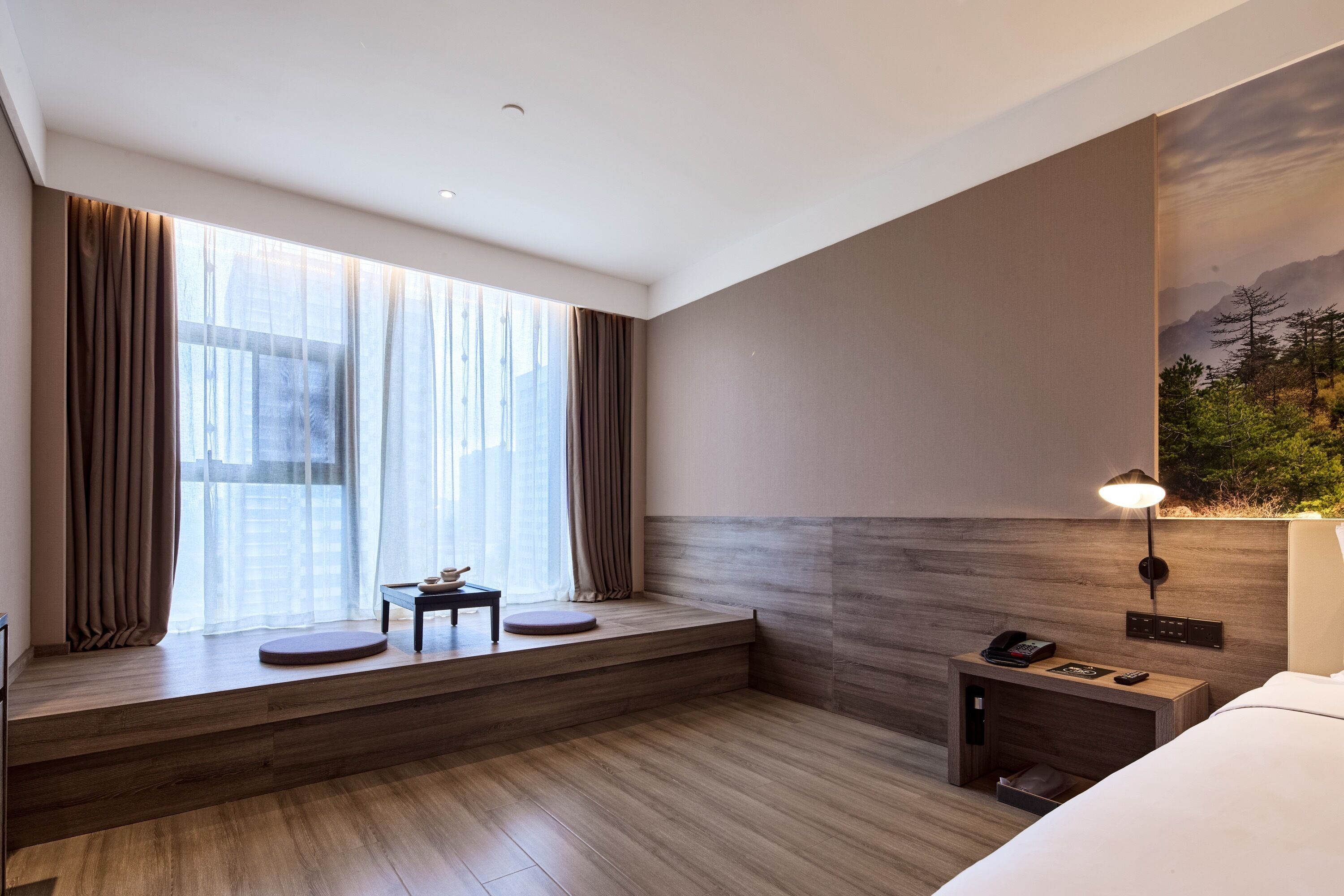 A modern hotel room with a large window, a wooden bench, a bedside table with a lamp, and a bed.