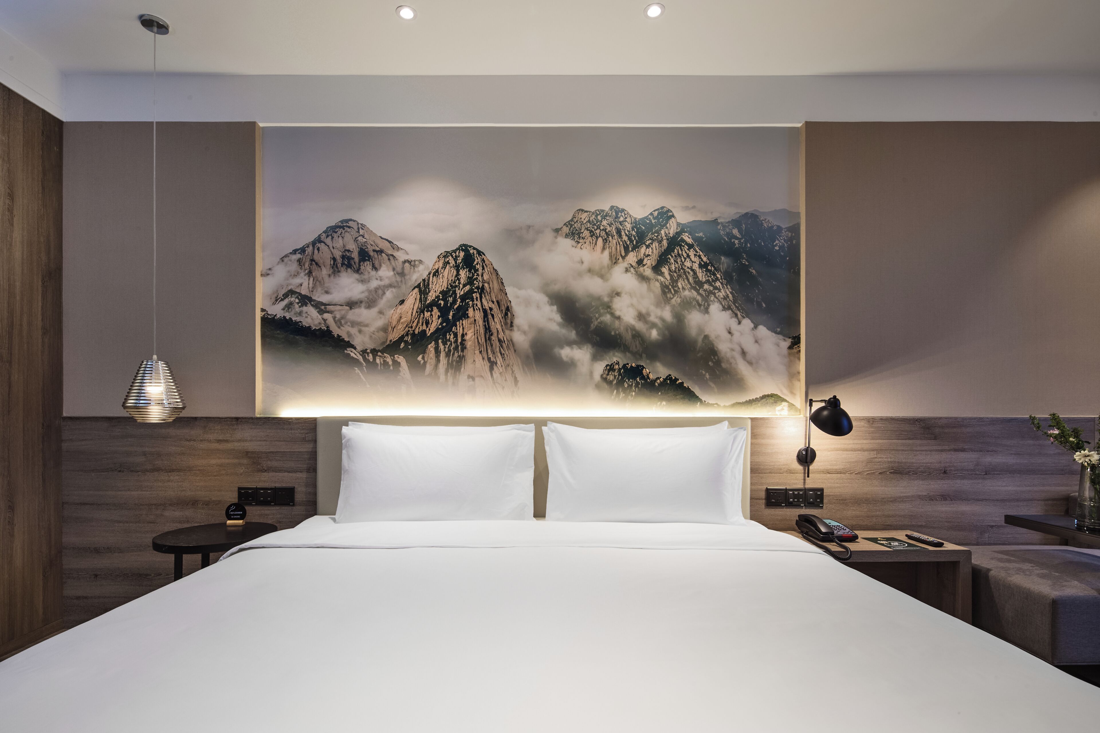 Una habitación de hotel moderna con una cama grande, una mesita de noche con lámpara, un teléfono en la pared y un cuadro decorativo de montañas.