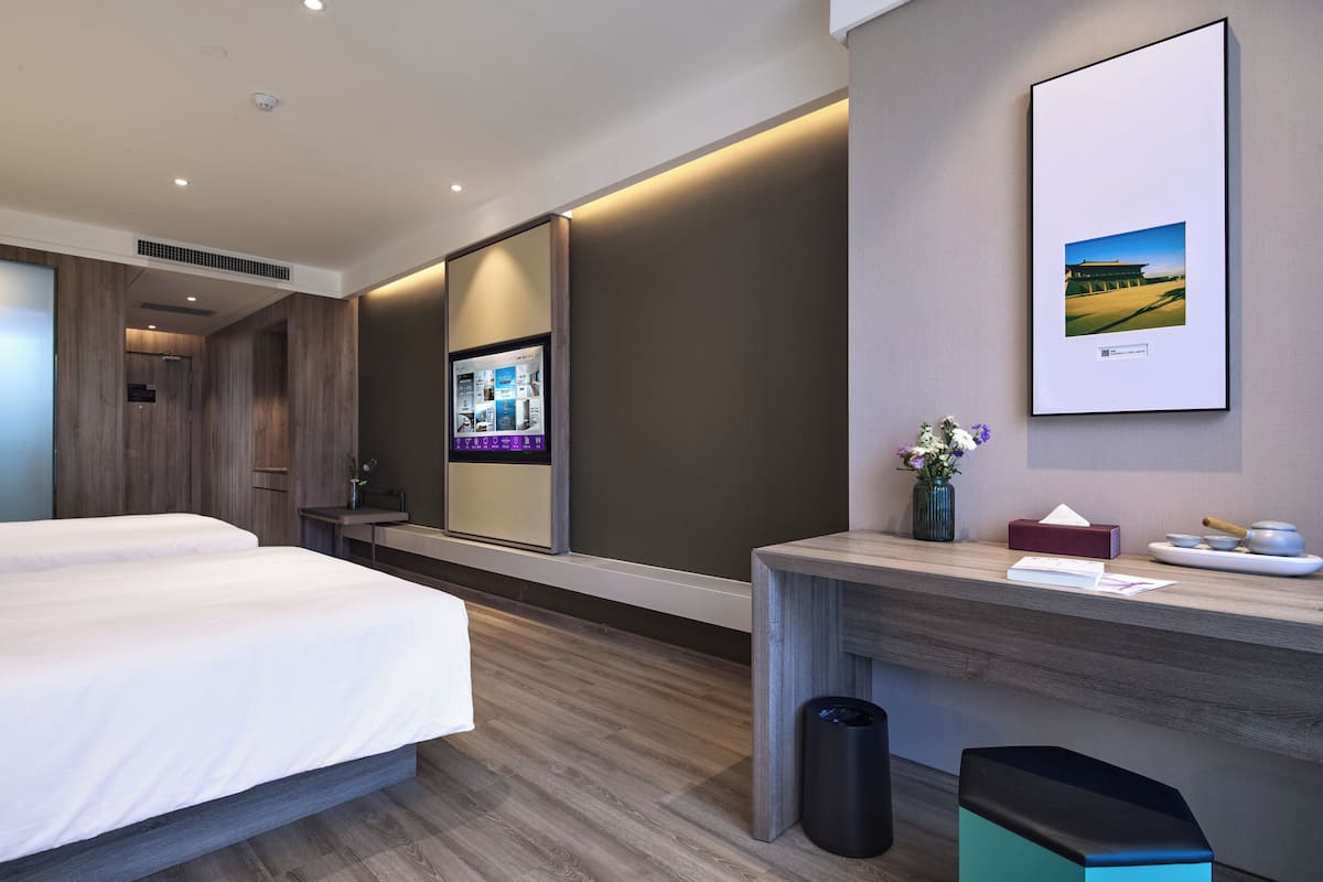 A hotel room with two beds, a desk, a trash bin, a framed picture, and a wall-mounted TV.