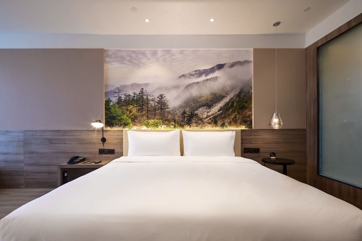 A modern hotel room with a large bed, a wall-mounted TV, and a scenic mountain landscape mural.