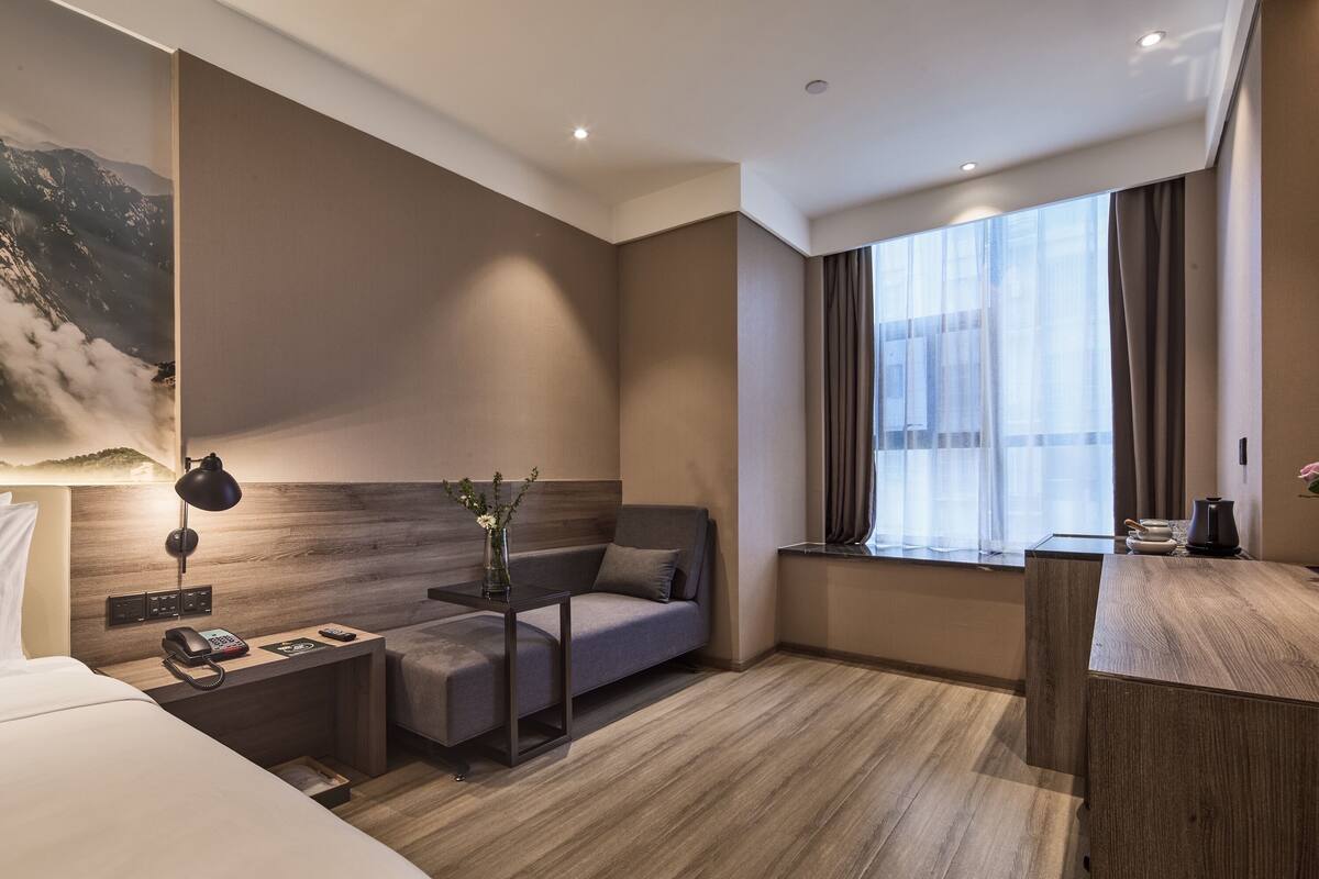 A modern hotel room with a wooden floor, a sofa bed, a small desk with a lamp, a phone, and a window with curtains.