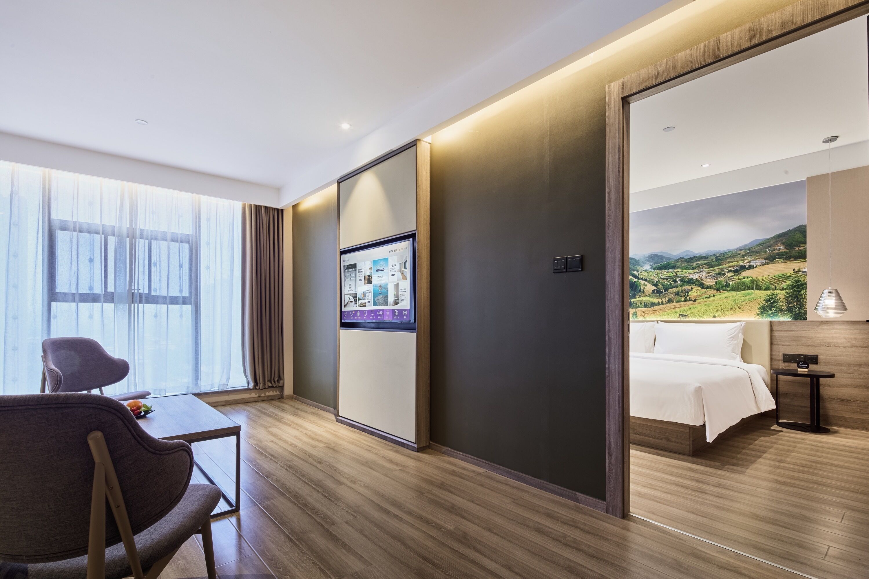 A modern hotel room with a wooden floor, a large window with sheer curtains, a flat-screen TV, a bed with white linens, and a small table with a lamp.