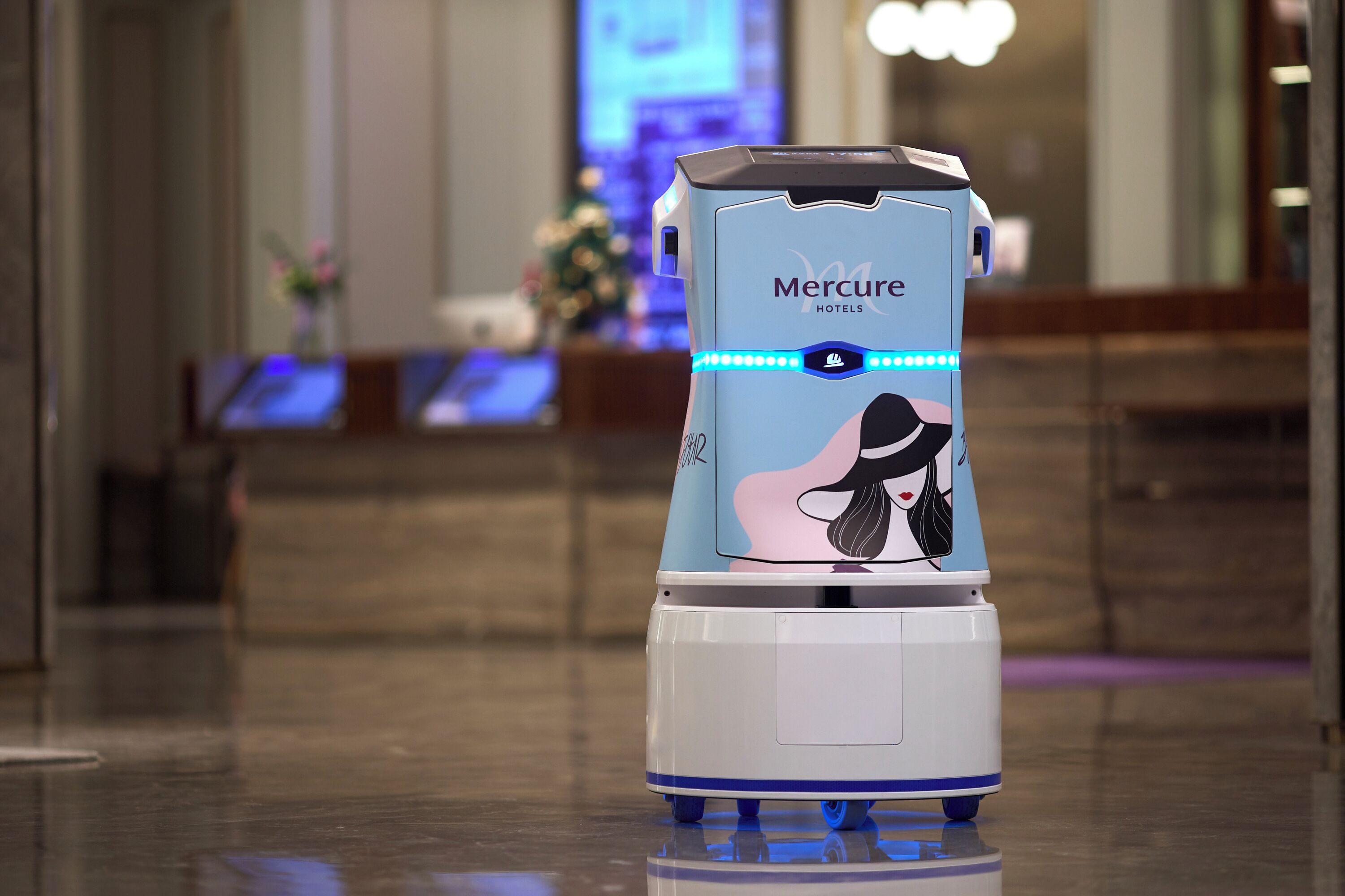 A robot with a Mercure Hotels logo, standing in a lobby with a reception desk and a blurred background.
