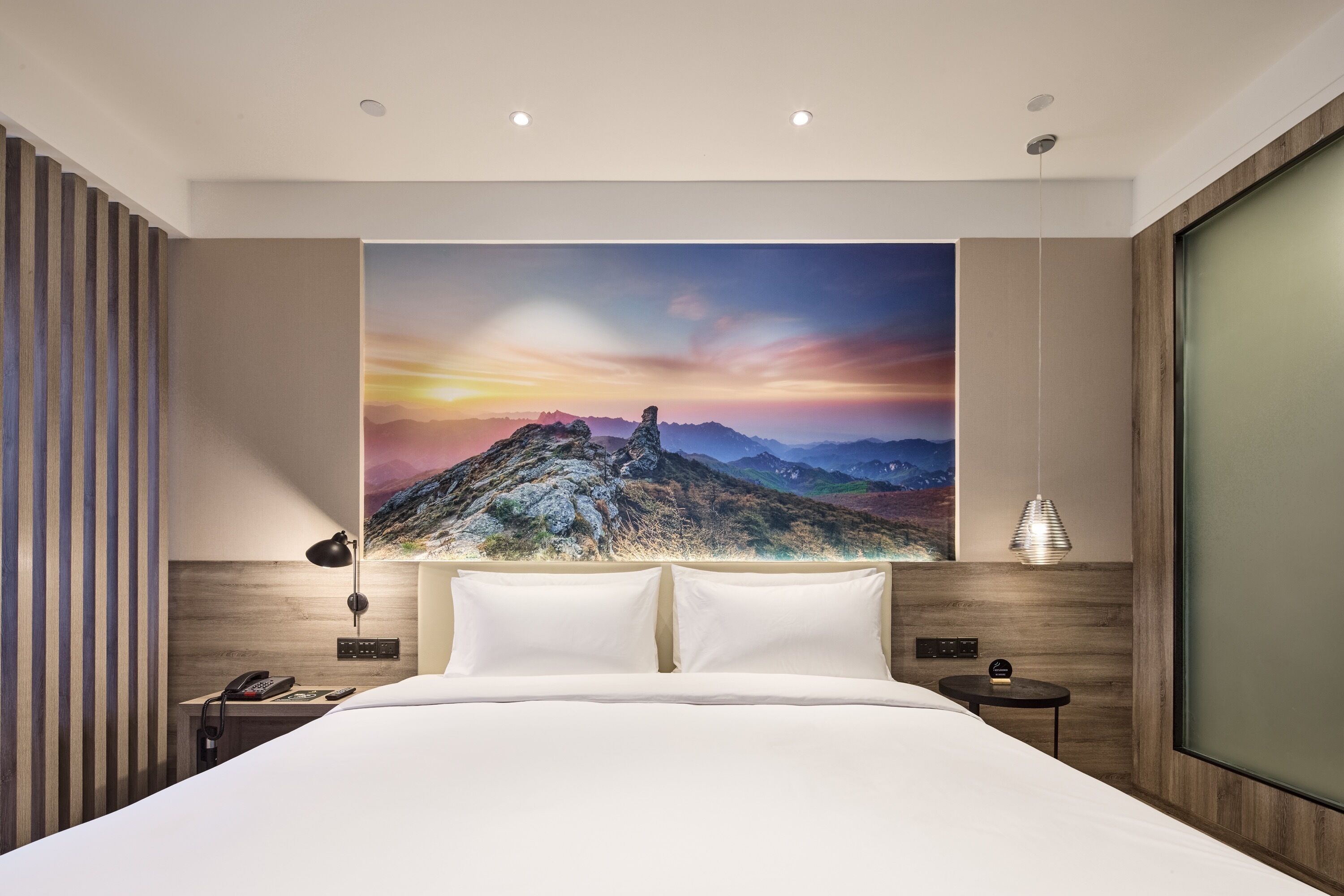 Une chambre d’hôtel moderne avec un grand lit, une table de chevet, une lampe et un tableau représentant un paysage de montagne accroché au mur.