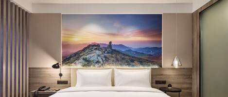 A modern hotel room with a large bed, a nightstand, a lamp, and a painting of a mountain landscape on the wall.
