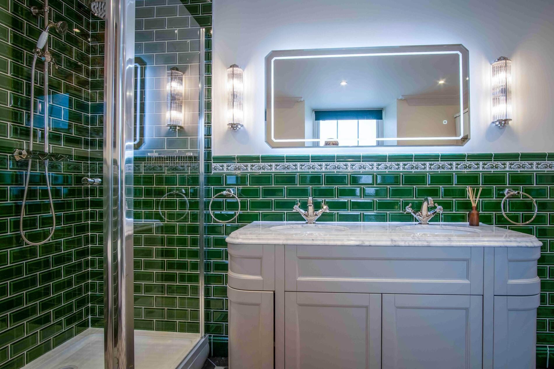 A bathroom with green mosaic tiles, a glass shower enclosure, a white vanity with a mirror, and wall-mounted lights.