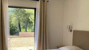 A bedroom with a bed, two pillows, a hanging light fixture, a window with curtains, and a view of trees and a fence. - Mini Villa, 15mn from Porto Vecchio (Sotta)