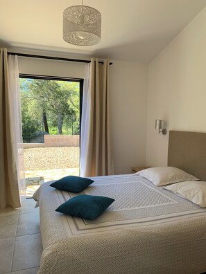 Una camera da letto con un letto, due cuscini, una lampada a sospensione, una finestra con tende e vista su alberi e una recinzione.