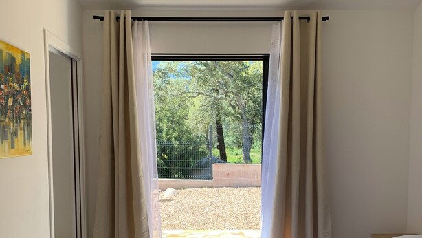 A bedroom with a bed, two pillows, a hanging light fixture, a window with curtains, and a view of a garden. - Mini Villa, 15mn from Porto Vecchio (Sotta)