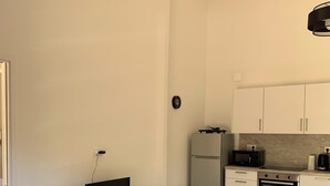 A modern kitchen with a built-in oven, microwave, and refrigerator. There's a flat-screen TV on a grey console table, a small black speaker, and a blue bowl. A glass table is in the foreground. - Mini Villa, 15mn from Porto Vecchio (Sotta)