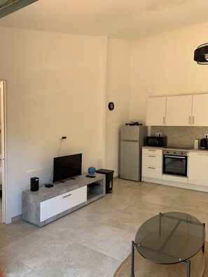 Una cucina moderna con forno, microonde e frigorifero integrati. C'è una TV a schermo piatto su un tavolino grigio, un piccolo altoparlante nero e una ciotola blu. Un tavolo in vetro è in primo piano.