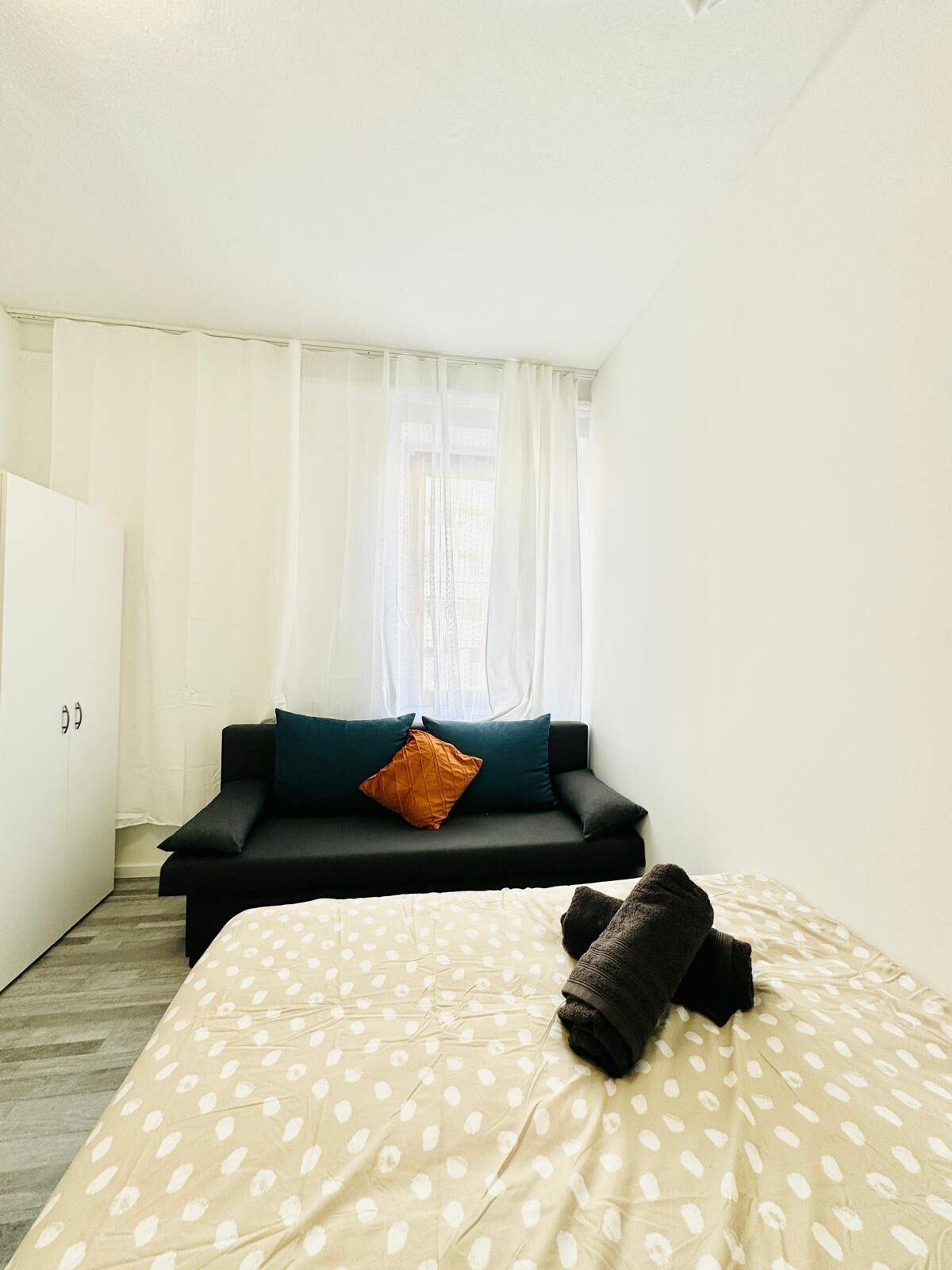 A minimalist room with a bed, a dark sofa, and a window with sheer curtains.
