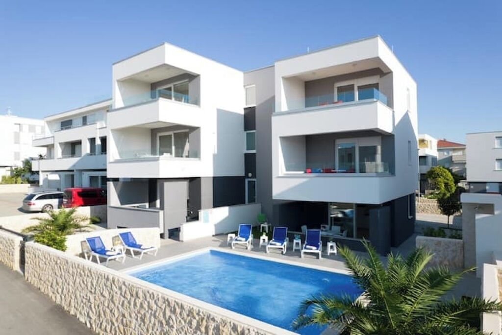 Un hotel moderno de varios pisos con piscina, sillas de descanso y un muro de piedra.