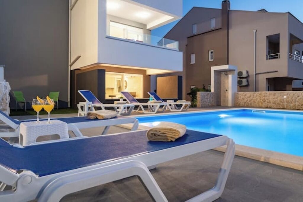 A modern outdoor pool area with lounge chairs and a table, a well-lit interior space, and a stone wall.