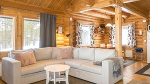 A log cabin interior with a light-colored sectional sofa, a small round table, and a dining area with a chair and table. - Kultaranta ii by Interhome (Inari)