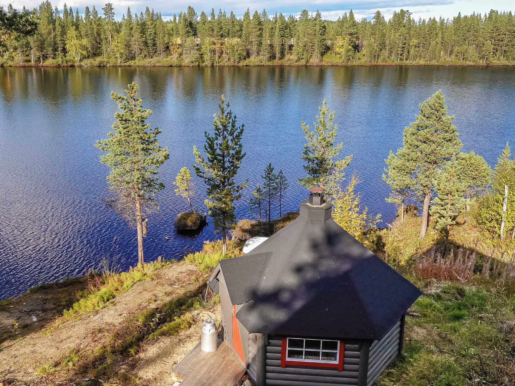 Une cabane avec un cadre de fenêtre rouge, entourée d’une forêt et d’un lac.