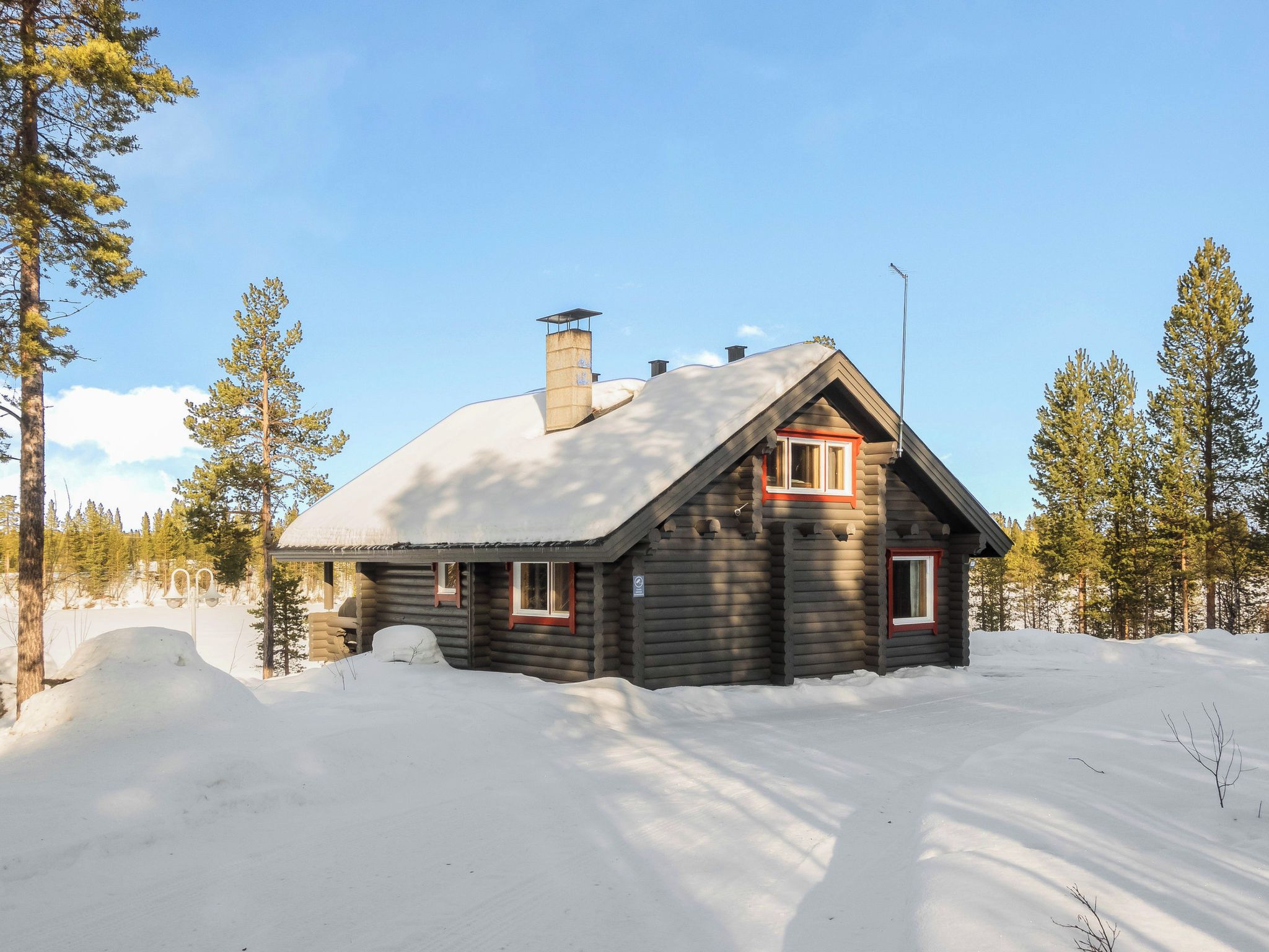 Lumihunnun peittämä hirsimökki, jonka ympärillä on lunta ja puita.