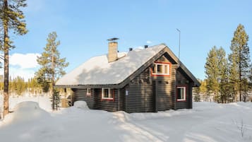 Lumihunnun peittämä hirsimökki, jonka ympärillä on lunta ja puita.