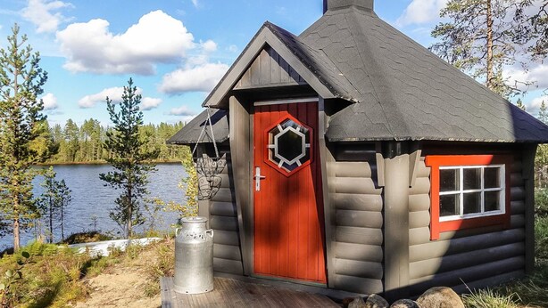 A log cabin with a red door and a hexagonal window, situated by a lake with trees in the background. - Kultaranta ii by Interhome (Inari)
