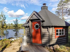 A log cabin with a red door and a hexagonal window, situated by a lake with trees in the background. - Kultaranta ii by Interhome (Inari)