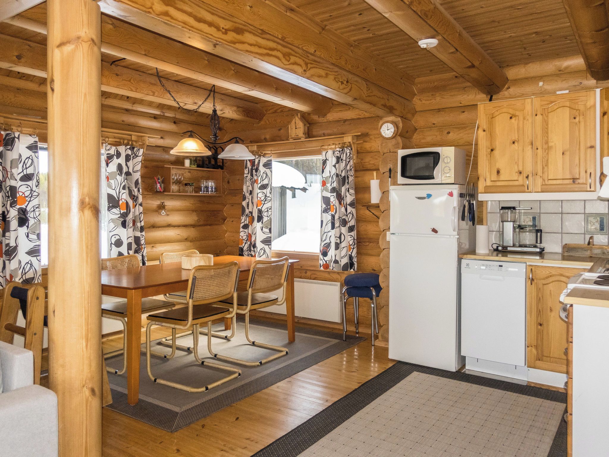 A wooden cabin interior with a dining area, kitchen, and a window with curtains.