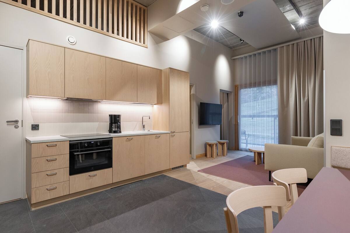 A modern kitchen with wooden cabinets, a built-in oven, and a coffee maker. There is a dining area with a table and chairs, and a large window with curtains.