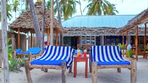 Un espace de détente aménagé au bord de la plage, avec des chaises longues rayées de bleu et de blanc, une petite table en bois sur laquelle sont posées des boissons, et des structures à toits de chaume.