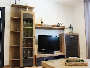 Een woonkamer met een flatscreen televisie, houten planken met kristal, een kleine tafel met decoratieve objecten en een kast.