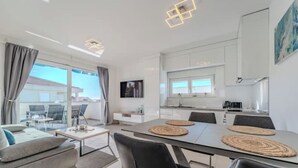 A modern, well-lit room with a dining table, chairs, and a sofa. There is a kitchen area with cabinets and a window. - Nova Excellence apartment (Novalja)