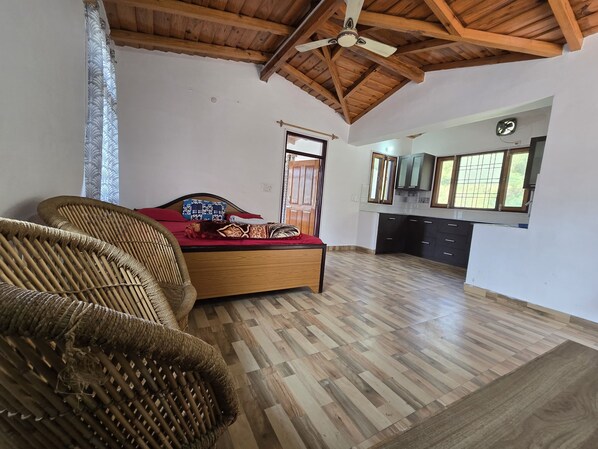 A room with a bed, a wicker chair, a wooden ceiling, and a view to a kitchen area.