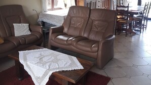 A living room with a brown leather sofa, a wooden coffee table with a white lace table runner, and a red rug. There is a window with curtains, a chandelier, and a dining area in the background. - Vacation home Bornem, quiet location. Close to Ghent, Antwerp, Mechelen, Puurs (Bornem)