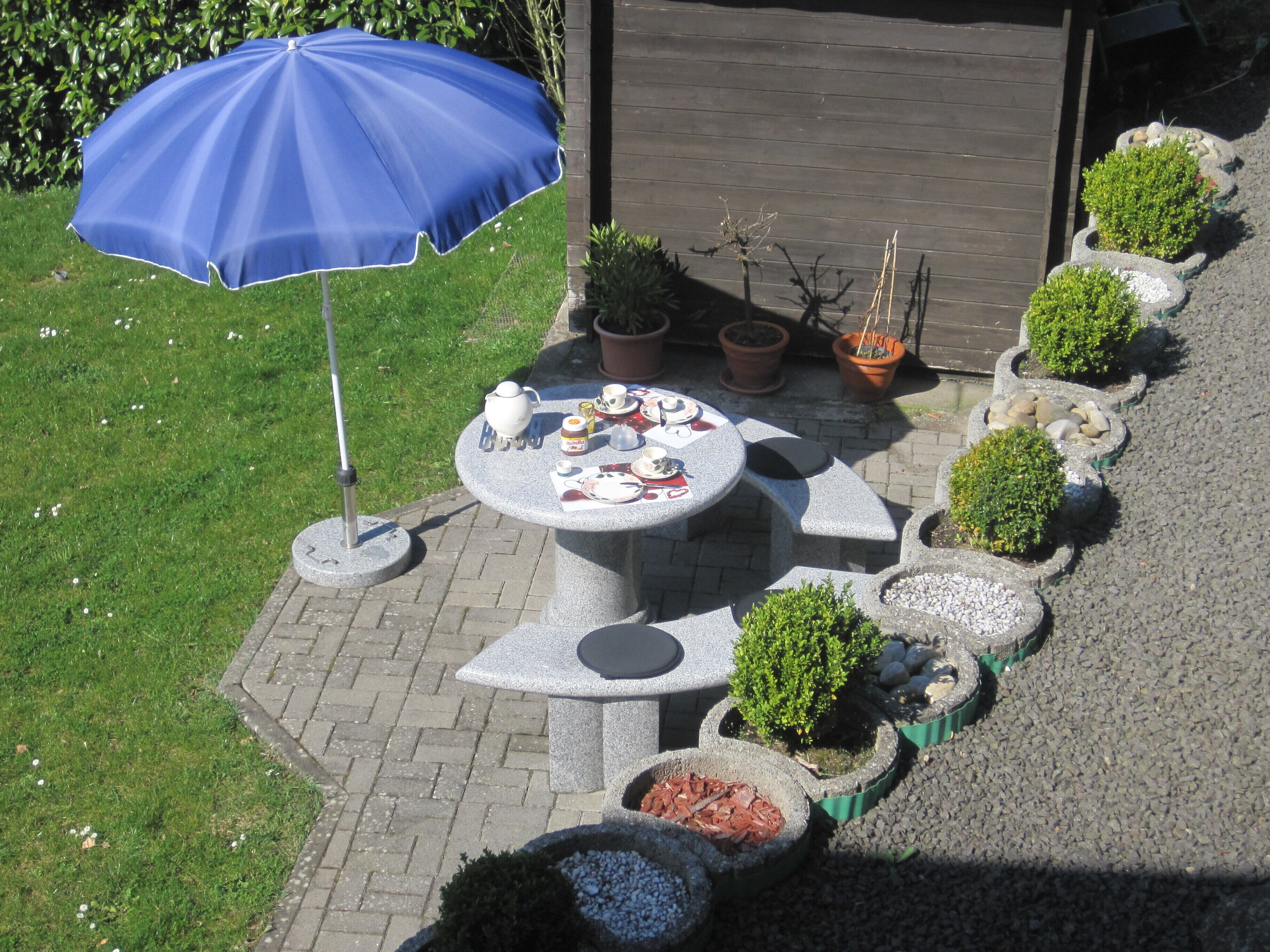 A patio with a round table set for tea, surrounded by potted plants and a blue umbrella.