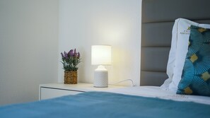 A neatly made bed with a blue blanket, a white bedside table with a lamp and a woven basket containing purple flowers, and a pillow with a geometric pattern. - Apartment Ouravale - By D´alma (Albufeira)
